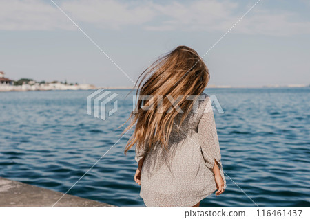 A woman is walking on the beach wearing a dress. The water is calm and the sky is clear. A woman is walking on the beach wearing a dress. The water is calm and the sky is clear. 116461437