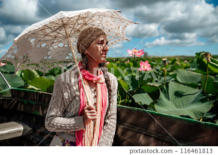 A woman wearing a hat and holding an umbrella is looking at a field of pink flowers. The scene is peaceful and serene, with the woman taking in the beauty of the flowers. 116461813
