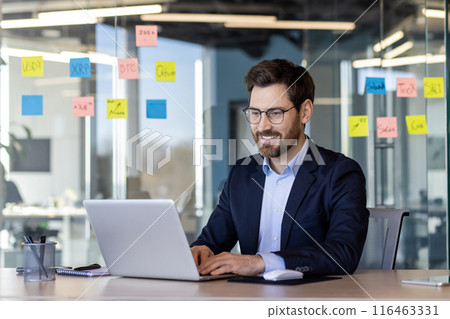 Successful smiling man working with laptop inside office, satisfied with results businessman typing on keyboard, mature boss in business suit at workplace. 116463331