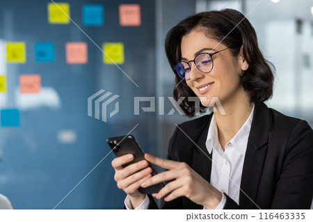 Confident businesswoman wearing glasses holding smartphone in modern office. Thoughtful professional checking messages on phone surrounded by sticky notes. Evening working atmosphere 116463335