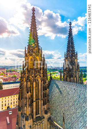 St. Vitus Cathedral ornate spires tower over the surrounding landscape with city and greenery in the backdrop under a partly cloudy sky. St. Vitus Cathedral ornate spires tower over the surrounding landscape with city and greenery in the backdrop under a partly cloudy sky. 116463341