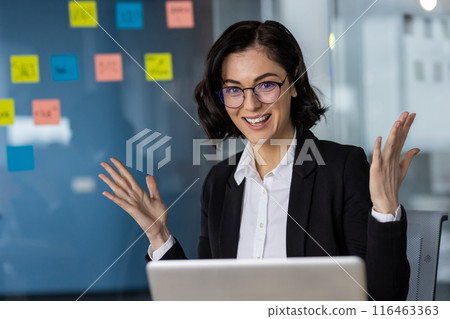 Confident businesswoman in a modern office expressing excitement during an online meeting. Woman wearing glasses and formal suit, gesturing enthusiastically while using laptop. 116463363