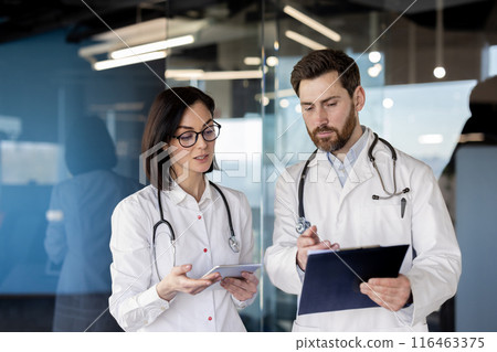 Two doctors in white coats reviewing patient data on a clipboard and tablet in contemporary medical office. Focused healthcare professionals collaborating during work meeting Two doctors in white coats reviewing patient data on a clipboard and tablet in contemporary medical office. Focused healthcare professionals collaborating during work meeting 116463375