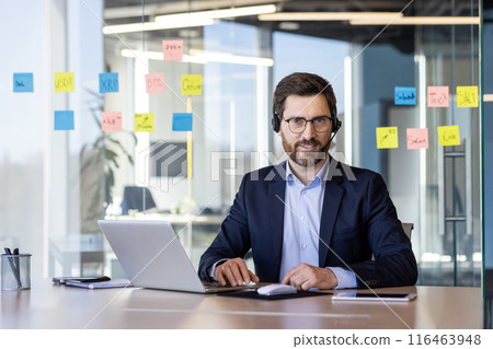 Portrait of successful businessman with headset phone, man in business suit smiling and looking at camera inside office, worker with laptop, helping buyers consumers online, support service. Portrait of successful businessman with headset phone, man in business suit smiling and looking at camera inside office, worker with laptop, helping buyers consumers online, support service. 116463948