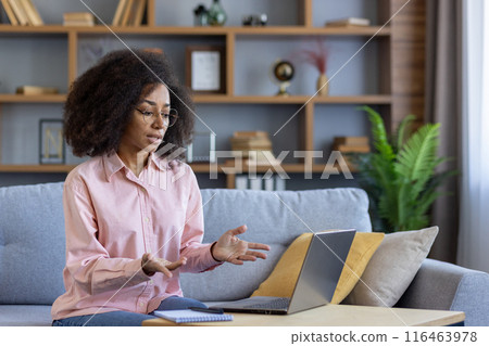 Woman sitting on couch engaging in virtual meeting on laptop. Working from home in casual attire. Bookshelves in background 116463978
