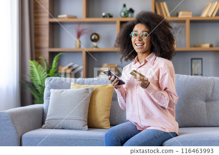 Smiling woman using smartphone and credit card for online shopping while sitting on cozy sofa 116463993