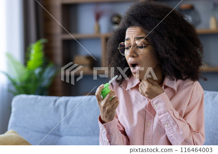 Woman in glasses using throat spray for sore throat relief while sitting on couch in living room 116464003