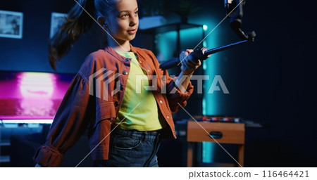 Energetic young girl using phone on selfie stick to film dance tutorial for other children watching her online social media content. Cheerful influencer recording video targeting generation Z fans 116464421