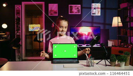 Cheerful child in home studio presenting advertisement from sponsoring brand on green screen laptop. Gen Z girl using chroma key device to present promotion from media partner to followers 116464451