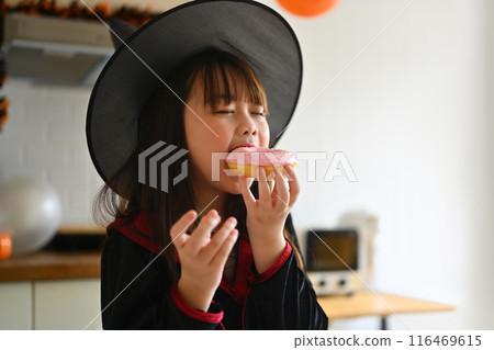 Satisfied pretty little girl in witch costumes eating delicious donut during Halloween celebration 116469615