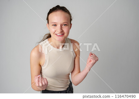 Young Woman in Casual Attire Smiling and Making a Fist Pump Gesture on a Plain Background Young Woman in Casual Attire Smiling and Making a Fist Pump Gesture on a Plain Background 116470490