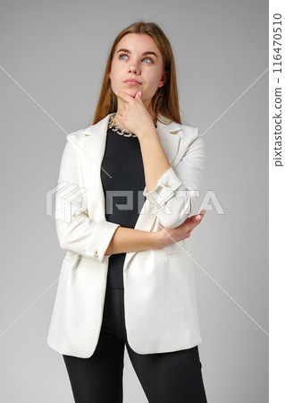 Thoughtful Young Woman in a Stylish White Blazer Posing on gray background 116470510