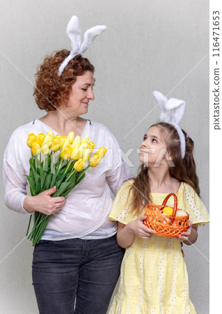 The family celebrates Easter. A woman with flowers and a girl with bunny ears and a basket of Easter eggs look at each other. 116471653