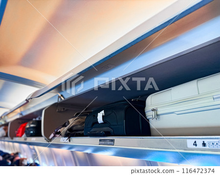 Overhead storage compartment inside airplane cabin filled with various carry on luggage, including black and white suitcases, backpacks, bags, with compartment doors open, visible seat row numbers. 116472374