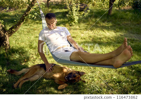 a man rests and listens to music in a hammock, his dog rests nearby 116473862