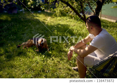 man rests and listens to music in a hammock, his dog rests nearby 116473872