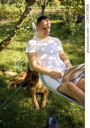 man works on a laptop in a hammock in the garden, next to his pet dog man works on a laptop in a hammock in the garden, next to his pet dog 116473906