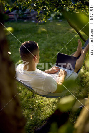 man works on a laptop in a hammock in the garden, next to his pet dog man works on a laptop in a hammock in the garden, next to his pet dog 116473912