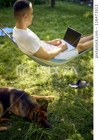 man works on a laptop in a hammock in the garden, next to his pet dog man works on a laptop in a hammock in the garden, next to his pet dog 116473913