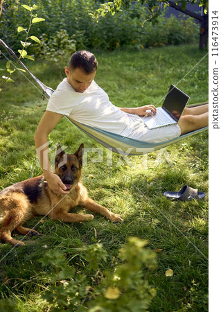 man works on a laptop in a hammock in the garden, next to his pet dog man works on a laptop in a hammock in the garden, next to his pet dog 116473914