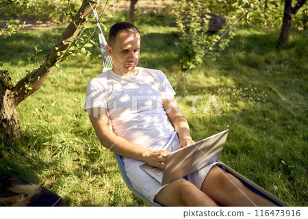 man works on a laptop in a hammock in the garden, next to his pet dog 116473916