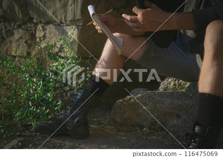 Male legs in old leather boots and shorts close up. A traveler with a notebook at a rest stop sits on a stone in an ancient fortress Male legs in old leather boots and shorts close up. A traveler with a notebook at a rest stop sits on a stone in an ancient fortress 116475069