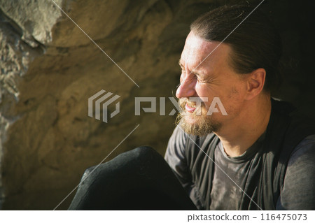Emotional Portrait close up. A man dressed as a traveler with a hat in his hand against the backdrop of an arched window of the fortress wall squints his eyes in the rays of the sunset. Archeology Emotional Portrait close up. A man dressed as a traveler with a hat in his hand against the backdrop of an arched window of the fortress wall squints his eyes in the rays of the sunset. Archeology 116475073
