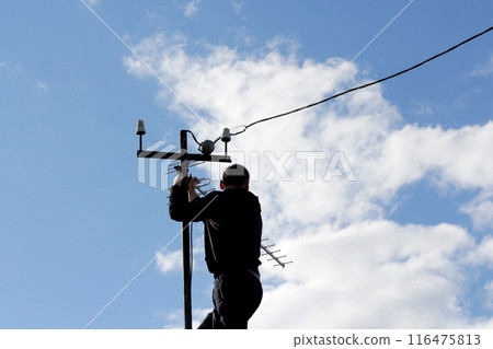 Master technician installs television antenna on rooftop. Expert setting up communication antenna under clear sky. Skilled technician mounting TV antenna with blue sky background Master technician installs television antenna on rooftop. Expert setting up communication antenna under clear sky. Skilled technician mounting TV antenna with blue sky background 116475813