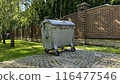 Metal trash can on wheels with lid against the backdrop of private household, trash container stands on the tiles near brick fence 116477546