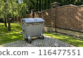 Trash on private territory against the backdrop of green lawn and brick fence, mobile trash container with lid near the house 116477551
