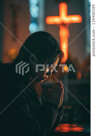 Christian woman stands in dimly lit room with white cross casting red glow on her face. She prays with hands clasped together, gazing upwards towards the cross. Quiet contemplation moment captured. 116480389