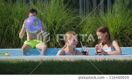 Two teenage girls enjoying drinks in a backyard pool while a man inflates a beach ball. 116480609
