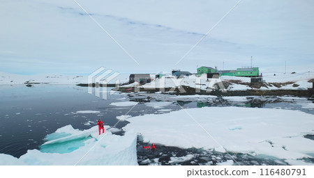 Scientists explore melting glaciers in Antarctica. Two man in orange wetsuits take samples standing on melt ice floe ocean water, climate change problem. Polar station expedition. Aerial drone flight 116480791