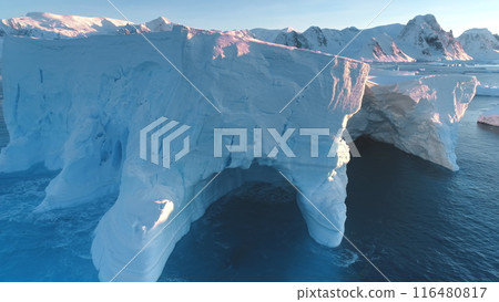 Huge iceberg towering in sunset light in Antarctica. Melting glacier holes and caves washed by ocean waves. Polar climate change at sunny winter day. Arctic winter landscape. Global warming problem. Huge iceberg towering in sunset light in Antarctica. Melting glacier holes and caves washed by ocean waves. Polar climate change at sunny winter day. Arctic winter landscape. Global warming problem. 116480817
