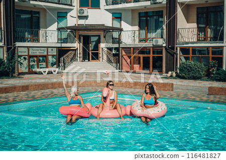 Three women are sitting on inflatable floats in a pool. Scene is lighthearted and fun. Three women are sitting on inflatable floats in a pool. Scene is lighthearted and fun. 116481827