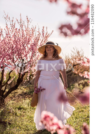 Woman blooming peach orchard. Against the backdrop of a picturesque peach orchard, a woman in a long white dress and hat enjoys a peaceful walk in the park, surrounded by the beauty of nature. Woman blooming peach orchard. Against the backdrop of a picturesque peach orchard, a woman in a long white dress and hat enjoys a peaceful walk in the park, surrounded by the beauty of nature. 116482090