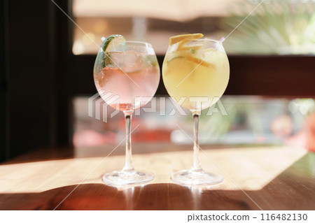 Two glasses of pink and yellow drinks sit on a table. The pink drink is a pink lemonade and the yellow drink is a lemonade with a slice of lime. 116482130