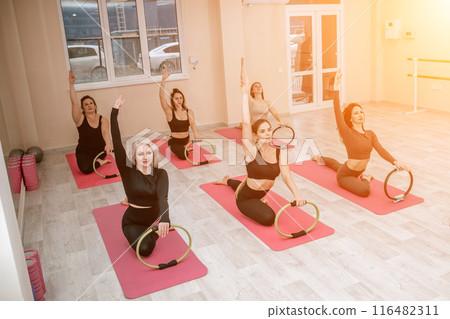 A group of six athletic women doing pilates or yoga on pink mats in front of a window in a beige loft studio interior. Teamwork, good mood and healthy lifestyle concept. A group of six athletic women doing pilates or yoga on pink mats in front of a window in a beige loft studio interior. Teamwork, good mood and healthy lifestyle concept. 116482311