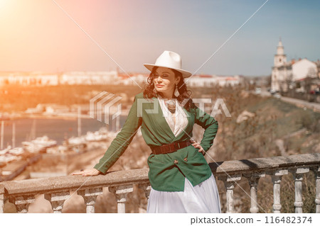 Woman walks around the city, lifestyle. Happy woman in a green jacket, white skirt and hat is sitting on a white fence with balusters overlooking the sea bay and the city. 116482374