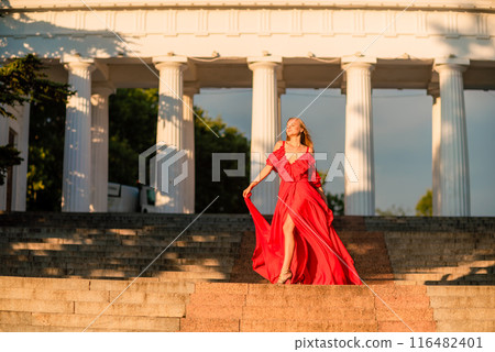 A woman in a long red dress against the backdrop of sunrise, bright golden light of the sun's rays. The concept of femininity, harmony. 116482401