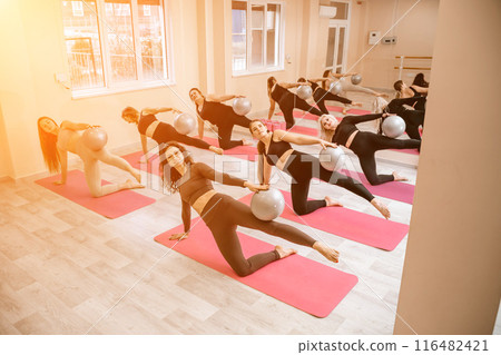 A group of six athletic women doing pilates or yoga on pink mats in front of a window in a beige loft studio interior. Teamwork, good mood and healthy lifestyle concept. A group of six athletic women doing pilates or yoga on pink mats in front of a window in a beige loft studio interior. Teamwork, good mood and healthy lifestyle concept. 116482421