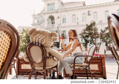 A woman sits cafe with a teddy bear next to her. The scene is set in a city with several chairs and tables around her. The woman is enjoying her time at the outdoor cafe. 116482431
