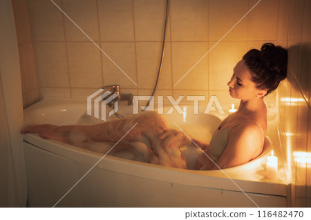 A woman is sitting in a bathtub with candles lit around her. Scene is relaxing and calming. A woman is sitting in a bathtub with candles lit around her. Scene is relaxing and calming. 116482470