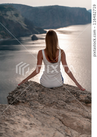 A woman sits on a rock overlooking the ocean. She is wearing a white tank top and white shorts. The scene is peaceful and serene, with the woman enjoying the view and the calming sound of the waves. A woman sits on a rock overlooking the ocean. She is wearing a white tank top and white shorts. The scene is peaceful and serene, with the woman enjoying the view and the calming sound of the waves. 116482719