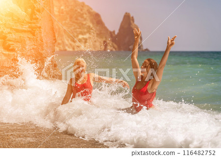 Women ocean play. Seaside, beach daytime, enjoying beach fun. Two women in red swimsuits enjoying themselves in the ocean waves and raising their hands up. 116482752