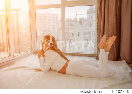 Side view portrait of relaxed woman listening to music with headphones lying on carpet at home. She is dressed in a white tracksuit. Side view portrait of relaxed woman listening to music with headphones lying on carpet at home. She is dressed in a white tracksuit. 116482758