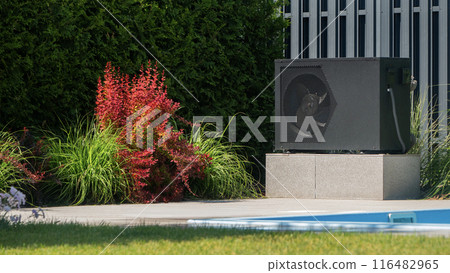 Outdoor unit of a heat pump at the edge of a pool. Decorative plants grow nearby. 116482965