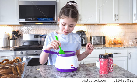 With focused attention, this budding culinary artist dips pretzels into a pot of melted chocolate, creating sweet delights in the warmth of a well-lit home kitchen. 116483883