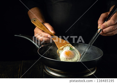 The cook cooks scrambled eggs in a frying pan and flips the egg onto the steaming frying pan using a kitchen spatula. Low key concept of preparing a delicious dinner 116485195
