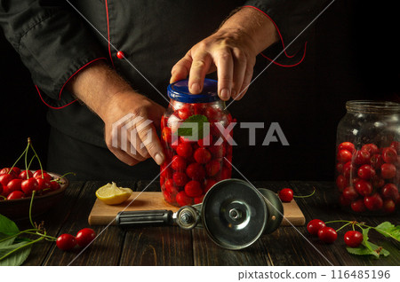 The cook closes a jar with a lid containing ripe cherries before rolling them up with a hand key. Making or preserving sweet cherry compote in the kitchen The cook closes a jar with a lid containing ripe cherries before rolling them up with a hand key. Making or preserving sweet cherry compote in the kitchen 116485196
